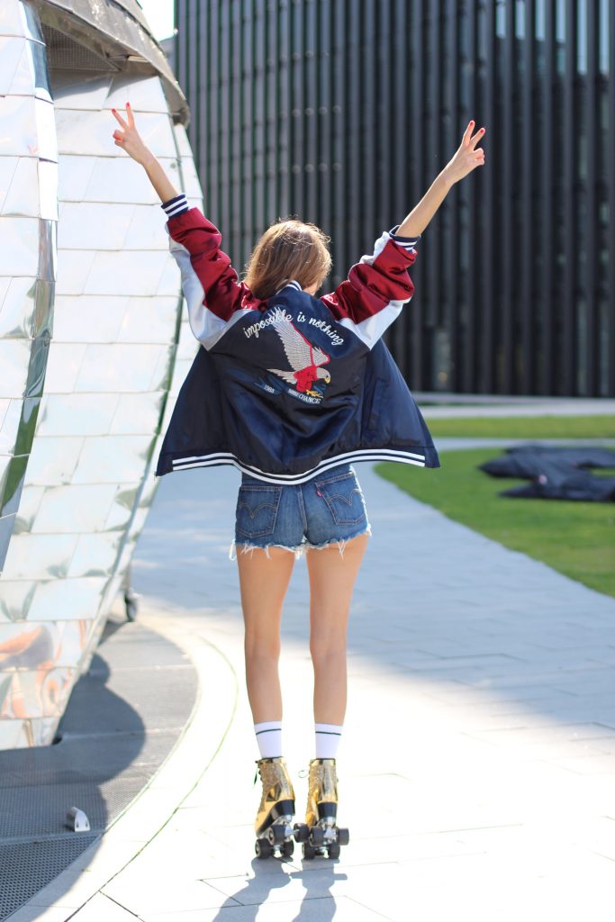 Model and Blogger Alexandra Lapp wearing golden, sparkling Roller Skates from Cosmoparis, mother-fucker socks from Mother Denim, Jadicted silk top in white, Levi's fringed denim high waist shorts, Stylenanda bomber jacket with an embroidered eagle and the letters 'Nothing is impossible' on the back of the three tone jacket in blue, white and red & Thom Browne aviator sunglasses in gold with red, in Duesseldorf on June, 2017.