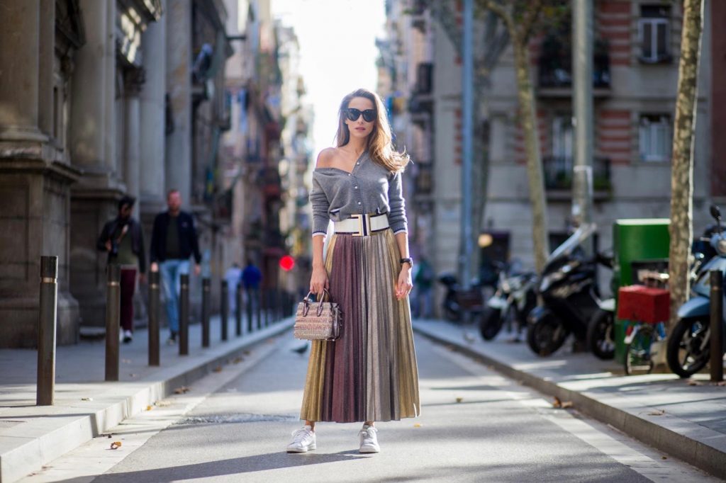 Alexandra Lapp wearing a long pleated metallic skirt with changing gold and red colors, grey cashmere cardigan with pearl buttons and blue and white rims, white and black lacquer waist belt from Balmain with gold buckle and details, Milla handbag in crystal tweed from MCM with metallic yarn and czech crystals, white and green Adidas Stan Smith sneakers and black Celine sunglasses on November 27, 2017 in Barcelona, Spain.