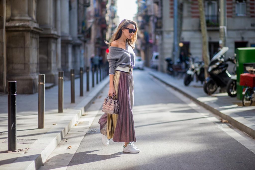 Alexandra Lapp wearing a long pleated metallic skirt with changing gold and red colors, grey cashmere cardigan with pearl buttons and blue and white rims, white and black lacquer waist belt from Balmain with gold buckle and details, Milla handbag in crystal tweed from MCM with metallic yarn and czech crystals, white and green Adidas Stan Smith sneakers and black Celine sunglasses on November 27, 2017 in Barcelona, Spain.