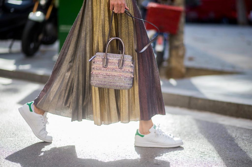 Alexandra Lapp wearing a long pleated metallic skirt with changing gold and red colors, grey cashmere cardigan with pearl buttons and blue and white rims, white and black lacquer waist belt from Balmain with gold buckle and details, Milla handbag in crystal tweed from MCM with metallic yarn and czech crystals, white and green Adidas Stan Smith sneakers and black Celine sunglasses on November 27, 2017 in Barcelona, Spain.