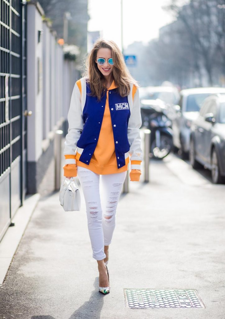 MILAN, ITALY - FEBRUARY 21: Alexandra Lapp wearing college style, a college jacket in blue, white and orange from MCM, an orange hoodie from glam-o-meter, white destroyed jeans from Frame a white Milla handbag from MCM, white pumps from Christian Louboutin and Ray-Ban sunglasses seen during Milan Fashion Week Fall/Winter 2018/19 on February 21, 2018 in Milan, Italy.
