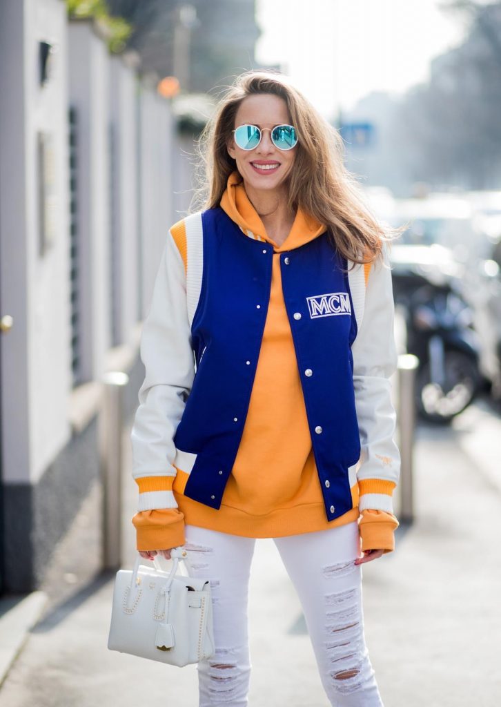 MILAN, ITALY - FEBRUARY 21: Alexandra Lapp wearing college style, a college jacket in blue, white and orange from MCM, an orange hoodie from glam-o-meter, white destroyed jeans from Frame a white Milla handbag from MCM, white pumps from Christian Louboutin and Ray-Ban sunglasses seen during Milan Fashion Week Fall/Winter 2018/19 on February 21, 2018 in Milan, Italy.