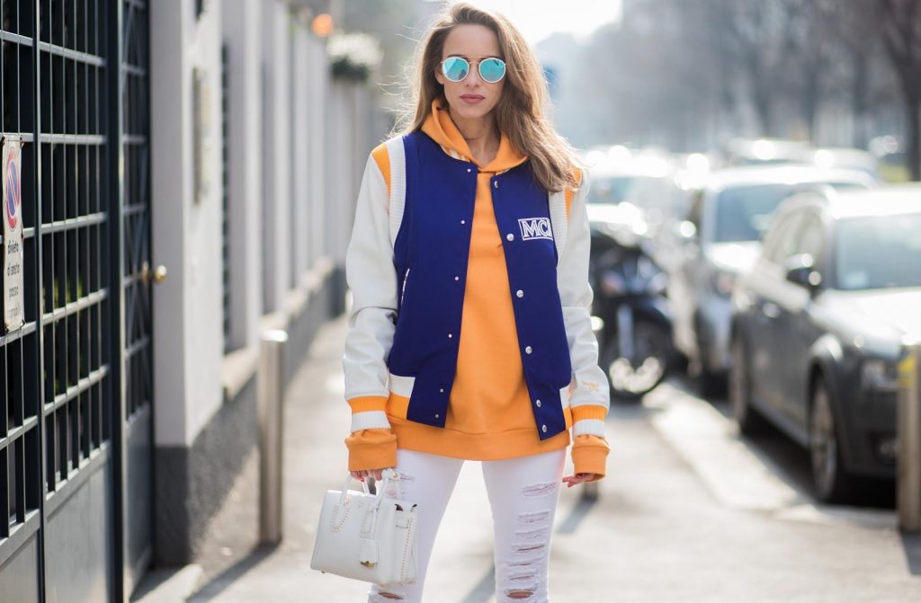 MILAN, ITALY - FEBRUARY 21: Alexandra Lapp wearing college style, a college jacket in blue, white and orange from MCM, an orange hoodie from glam-o-meter, white destroyed jeans from Frame a white Milla handbag from MCM, white pumps from Christian Louboutin and Ray-Ban sunglasses seen during Milan Fashion Week Fall/Winter 2018/19 on February 21, 2018 in Milan, Italy.