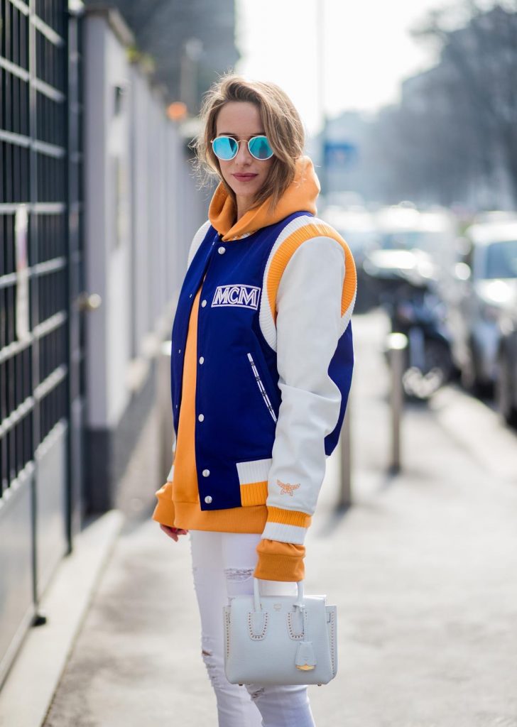 MILAN, ITALY - FEBRUARY 21: Alexandra Lapp wearing college style, a college jacket in blue, white and orange from MCM, an orange hoodie from glam-o-meter, white destroyed jeans from Frame a white Milla handbag from MCM, white pumps from Christian Louboutin and Ray-Ban sunglasses seen during Milan Fashion Week Fall/Winter 2018/19 on February 21, 2018 in Milan, Italy.