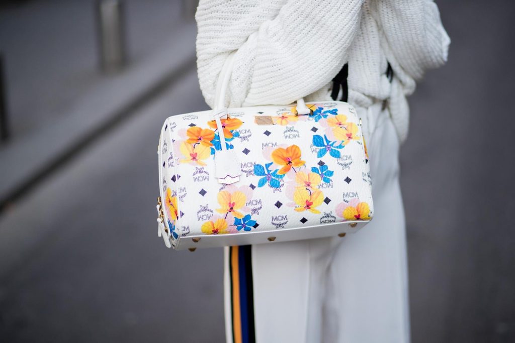 MCM, Zara, Chanel, Isabel Marant Alexandra Lapp wearing white sport pants with stripes on the side and a white knit v neck pullover from Zara, a white Baker Boy Cap from Chanel, suspenders from Chanel in white with black logo writing, black boots from Isabel Marant and Boston bag in floral print MCM is seen during Paris Fashion Week Womenswear Fall/Winter 2018/2019 on March 4, 2018 in Paris, France.