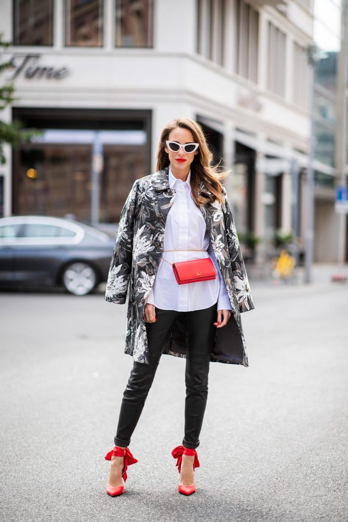 Alexandra Lapp in Floral Blazer Coat Look wearing a long blazer jacket with floral prints in green, white and gold tones from Steffen Schraut, a white long shirt and slim leather pants in black with stitches around the knee from Steffen Schraut, the red Douce Du Desert 100 Flamenco Satin sandals from Christian Louboutin with a long ribbon tied around the ankle, a red little clutch from Christian Louboutin around the waist and white Saint Laurent New Wave Lily sunglasses is seen during the Berlin Fashion Week July 2018 on July 6, 2018 in Berlin, Germany.