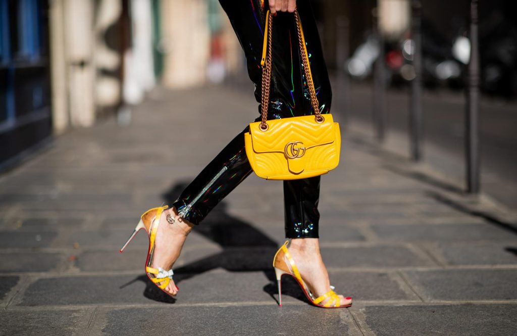 Alexandra Lapp in a Vinyl Pants Look wearing black zipped patent leather trousers from Balmain, a yellow v-neck knit jumper by Allude, shiny black down jacket by Ahirain, yellow Gucci Matelasse GG bag, yellow white Metrisandal open toe sandals with tape-style straps by Christian Louboutin and cat-eye shaped Isabella Sunglasses by illesteva during Paris Fashion Week Womenswear Spring/Summer 2019 on September 27, 2018 in Paris, France.