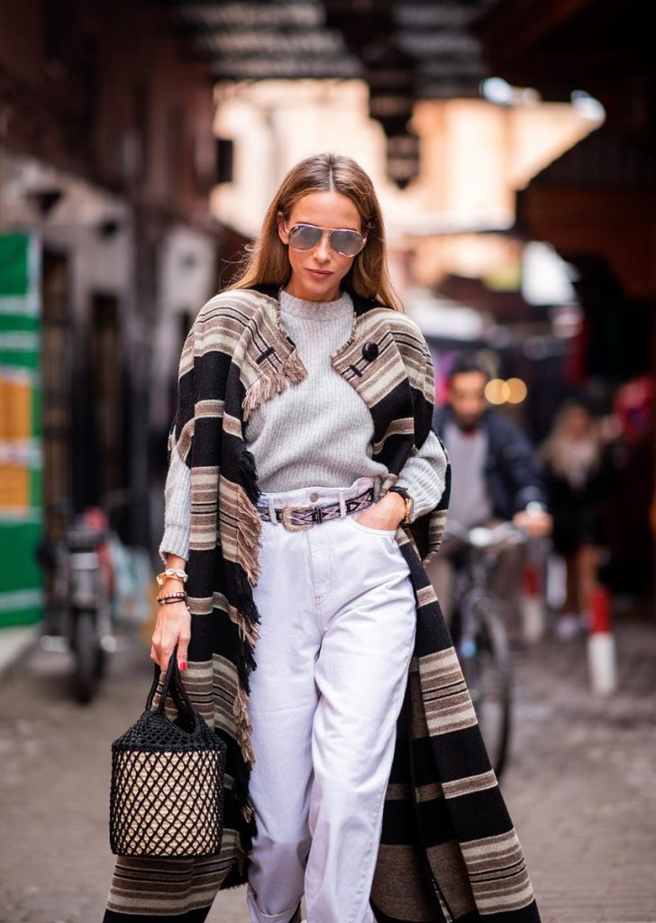 Alexandra Lapp in a Poncho Style wearing a long Huan fringed striped wool cape from Isabel Marant, an oversized grey wool sweater from H&M, white high-rise boyfriend style jeans from Isabel Marant Etoile, a black leather belt with white stitches from Isabel Marant, black Cowboy boots with a silver tip from Isabel Marant, a handwoven basket bag from Sensistudio and mirrored sunglasses from Givenchy on November 25, 2018 in Marrakech, Morocco.