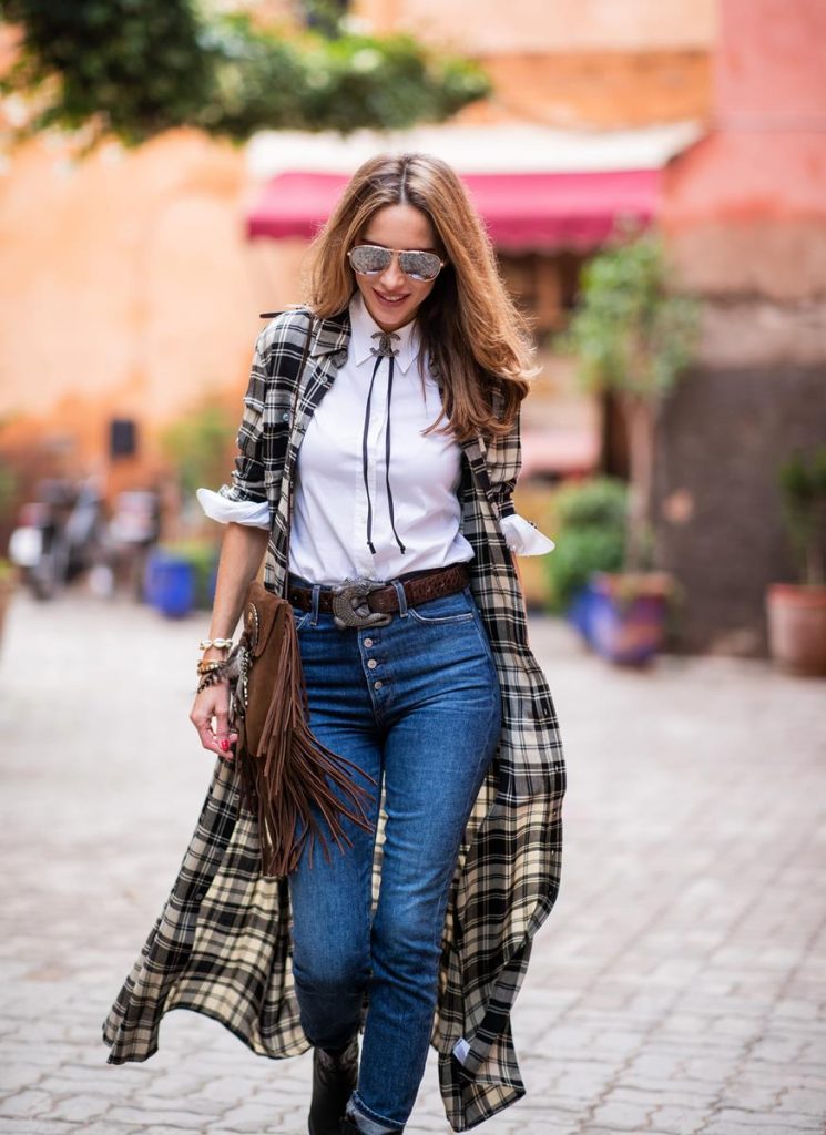 Polo Ralph Lauren, Loro Piana, Adriano Goldschmied, Saint Laurent, Isabel Marant, Christian Dior, Chanel, Givenchy Alexandra Lapp in a Cowboy Style Look wearing a long checkered shirt dress from Polo Ralph Lauren, a white shirt from Loro Piana, light blue denims from Adriano Goldschmied, a brown suede leather bag with long fringes and a buckle with feathers from Saint Laurent, a black leather belt with white stitches from Isabel Marant, black Cowboy boots from Christian Dior, a silver Chanel brooch and mirrored sunglasses from Givenchy on November 25, 2018 in Marrakech, Morocco.