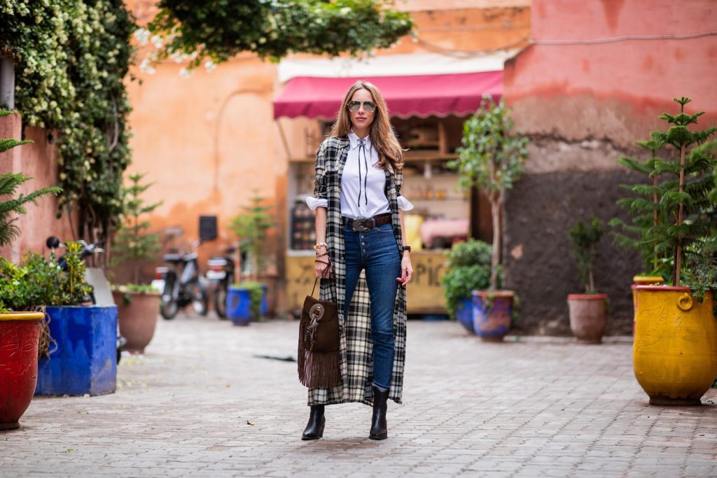 Polo Ralph Lauren, Loro Piana, Adriano Goldschmied, Saint Laurent, Isabel Marant, Christian Dior, Chanel, Givenchy Alexandra Lapp in a Cowboy Style Look wearing a long checkered shirt dress from Polo Ralph Lauren, a white shirt from Loro Piana, light blue denims from Adriano Goldschmied, a brown suede leather bag with long fringes and a buckle with feathers from Saint Laurent, a black leather belt with white stitches from Isabel Marant, black Cowboy boots from Christian Dior, a silver Chanel brooch and mirrored sunglasses from Givenchy on November 25, 2018 in Marrakech, Morocco.