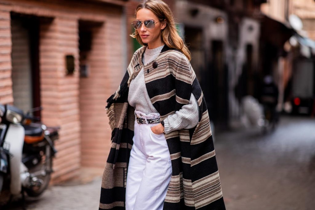 Alexandra Lapp in a Poncho Style wearing a long Huan fringed striped wool cape from Isabel Marant, an oversized grey wool sweater from H&M, white high-rise boyfriend style jeans from Isabel Marant Etoile, a black leather belt with white stitches from Isabel Marant, black Cowboy boots with a silver tip from Isabel Marant, a handwoven basket bag from Sensistudio and mirrored sunglasses from Givenchy on November 25, 2018 in Marrakech, Morocco.