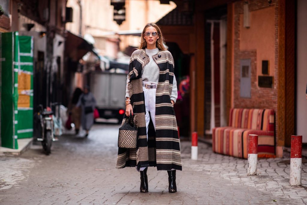 Alexandra Lapp in a Poncho Style wearing a long Huan fringed striped wool cape from Isabel Marant, an oversized grey wool sweater from H&M, white high-rise boyfriend style jeans from Isabel Marant Etoile, a black leather belt with white stitches from Isabel Marant, black Cowboy boots with a silver tip from Isabel Marant, a handwoven basket bag from Sensistudio and mirrored sunglasses from Givenchy on November 25, 2018 in Marrakech, Morocco.