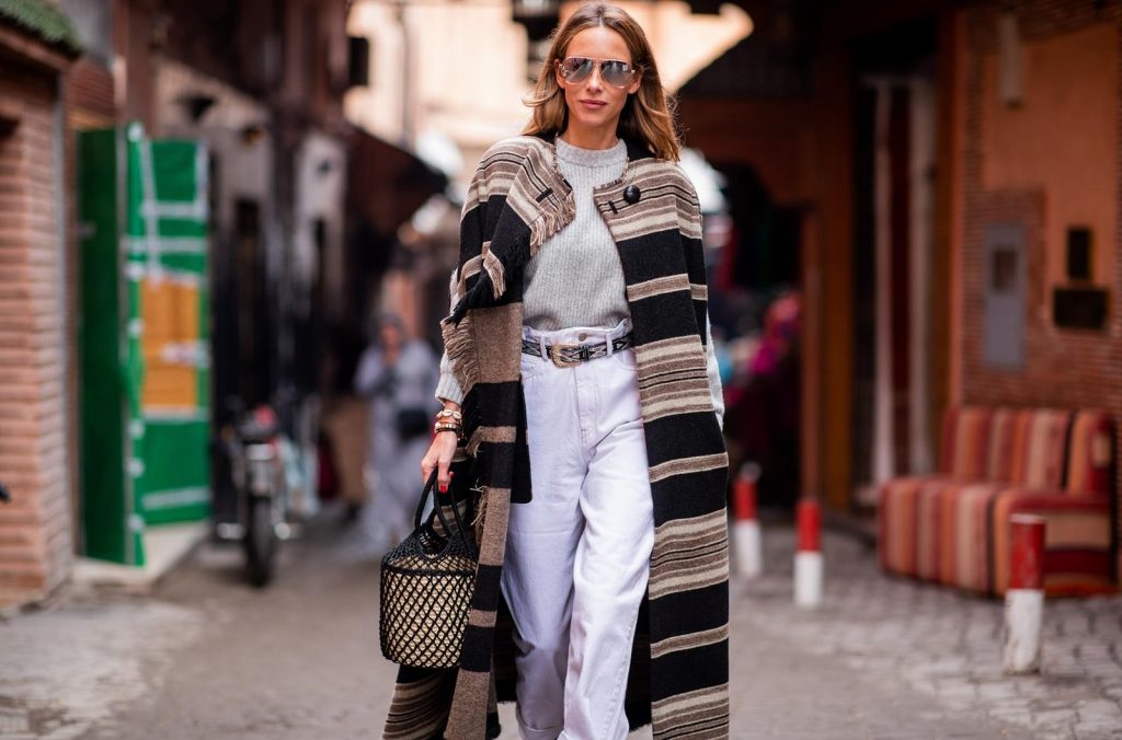 Alexandra Lapp in a Poncho Style wearing a long Huan fringed striped wool cape from Isabel Marant, an oversized grey wool sweater from H&M, white high-rise boyfriend style jeans from Isabel Marant Etoile, a black leather belt with white stitches from Isabel Marant, black Cowboy boots with a silver tip from Isabel Marant, a handwoven basket bag from Sensistudio and mirrored sunglasses from Givenchy on November 25, 2018 in Marrakech, Morocco.