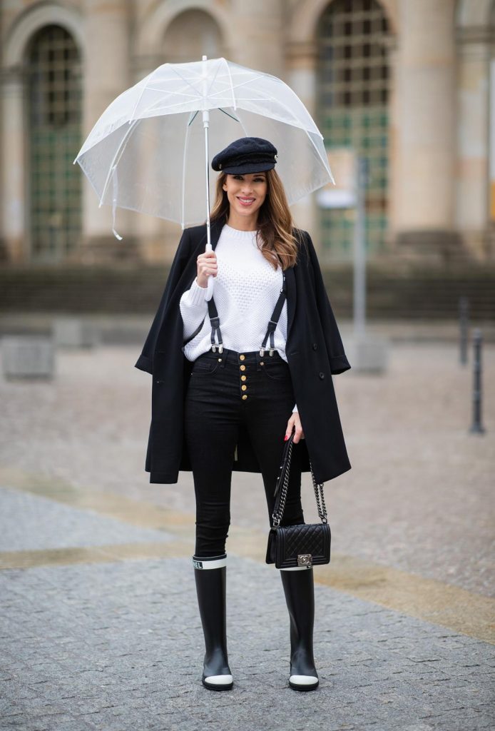 Alexandra Lapp in A Chanel Rain Boots look, wearing a soft long blazer by Steffen Schraut, over a white chunky knit sweater by H&M, black skinny pants with golden buttons by Veronica Beard, styled with Chanel logo suspenders and Chanel rain boots, the Chanel Le boy bag, a Chanel tweed baker boy cap and a transparent umbrella by Mirviory during the Berlin Fashion Week Autumn/Winter 2019 on January 15, 2019 in Berlin, Germany. (Photo by Christian Vierig/Getty Images)