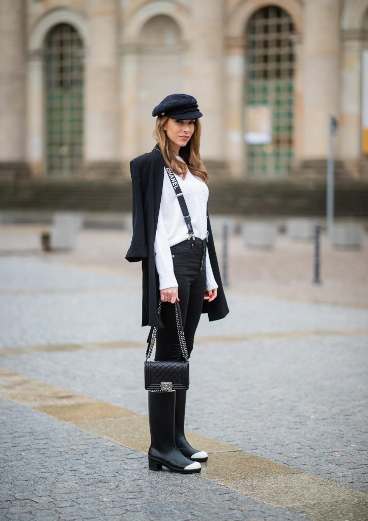 Alexandra Lapp in A Chanel Rain Boots look, wearing a soft long blazer by Steffen Schraut, over a white chunky knit sweater by H&M, black skinny pants with golden buttons by Veronica Beard, styled with Chanel logo suspenders and Chanel rain boots, the Chanel Le boy bag, a Chanel tweed baker boy cap and a transparent umbrella by Mirviory during the Berlin Fashion Week Autumn/Winter 2019 on January 15, 2019 in Berlin, Germany. (Photo by Christian Vierig/Getty Images)