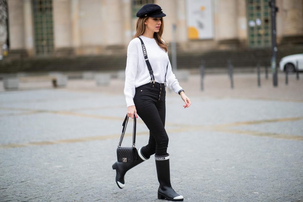 Alexandra Lapp in A Chanel Rain Boots look, wearing a soft long blazer by Steffen Schraut, over a white chunky knit sweater by H&M, black skinny pants with golden buttons by Veronica Beard, styled with Chanel logo suspenders and Chanel rain boots, the Chanel Le boy bag, a Chanel tweed baker boy cap and a transparent umbrella by Mirviory during the Berlin Fashion Week Autumn/Winter 2019 on January 15, 2019 in Berlin, Germany. (Photo by Christian Vierig/Getty Images)