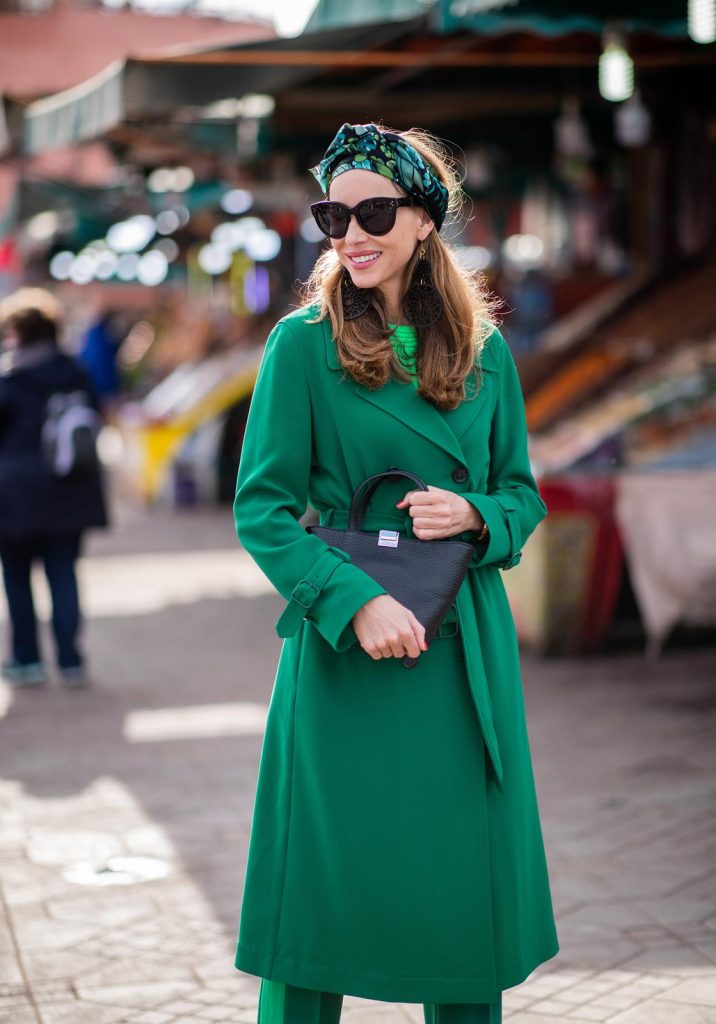 Alexandra Lapp in a Le Riad look, wearing a belted long crepe Marc Cain coat in green, a neon green sweater and crepe loose-fit pants with stripes in two green tones by Marc Cain, a black small trapeze shaped bag and a printed silk scarf in multi green shades by Marc Cain, black lacquer high heels, black sunglasses and round oversized earrings in black by Marc Cain on November 26, 2018 in Marrakech, Morocco. 