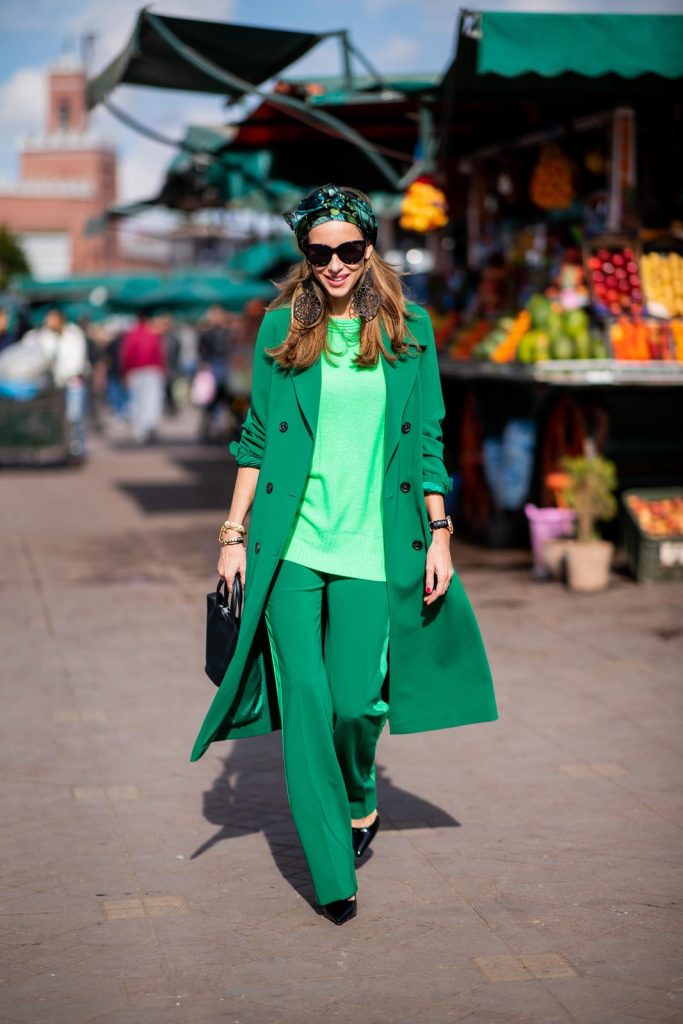 Alexandra Lapp in a Le Riad look, wearing a belted long crepe Marc Cain coat in green, a neon green sweater and crepe loose-fit pants with stripes in two green tones by Marc Cain, a black small trapeze shaped bag and a printed silk scarf in multi green shades by Marc Cain, black lacquer high heels, black sunglasses and round oversized earrings in black by Marc Cain on November 26, 2018 in Marrakech, Morocco. 