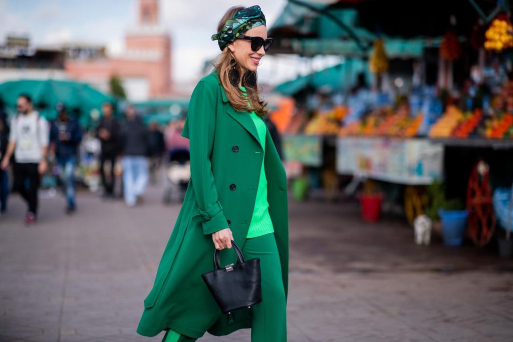 Alexandra Lapp in a Le Riad look, wearing a belted long crepe Marc Cain coat in green, a neon green sweater and crepe loose-fit pants with stripes in two green tones by Marc Cain, a black small trapeze shaped bag and a printed silk scarf in multi green shades by Marc Cain, black lacquer high heels, black sunglasses and round oversized earrings in black by Marc Cain on November 26, 2018 in Marrakech, Morocco. 