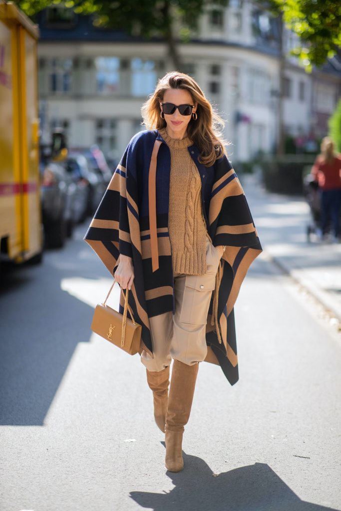 Alexandra Lapp is seen wearing Chloé striped wool cape, Chloé chunky-knit sleeveless jumper in camel, Chloé linen cargo pants in beige, SAINT LAURENT Kate medium bag in camel and CELINE Claude calfskin autumn boots in sand. All looks by Breuninger.