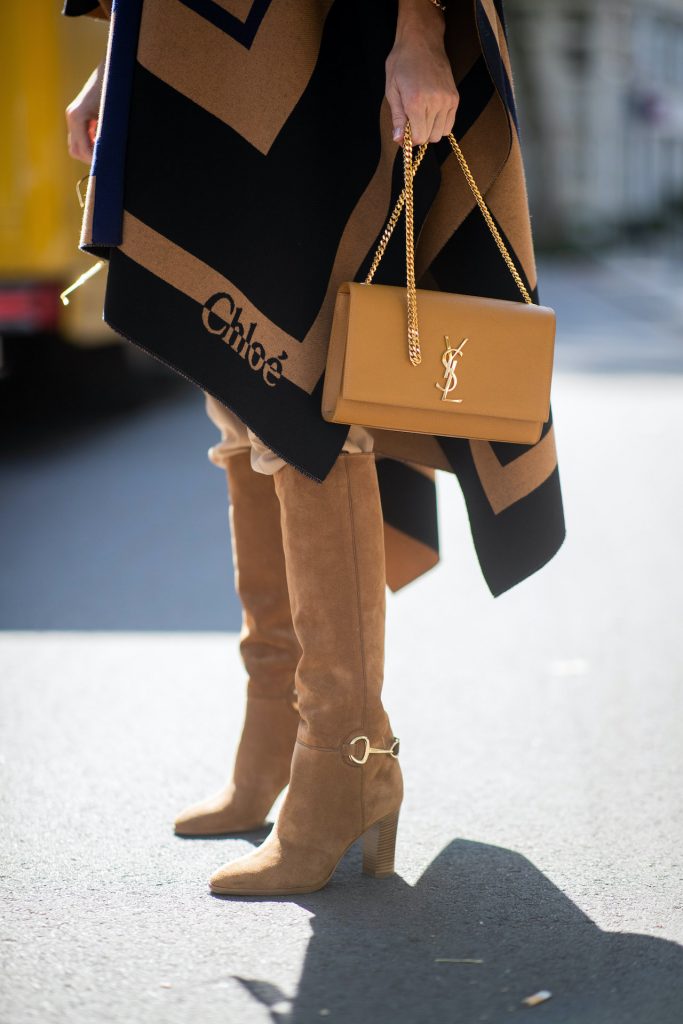 Alexandra Lapp is seen wearing Chloé striped wool cape, Chloé chunky-knit sleeveless jumper in camel, Chloé linen cargo pants in beige, SAINT LAURENT Kate medium bag in camel and CELINE Claude calfskin autumn boots in sand. All looks by Breuninger.